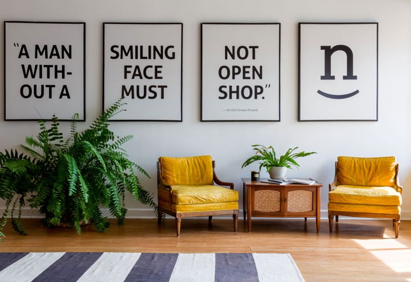 Beautiful space with a large fern, two yellow upholstered lounge chairs, a nicely decorated coffee table and four-panels of large wall art reading: A man without a smiling face must not open shop.