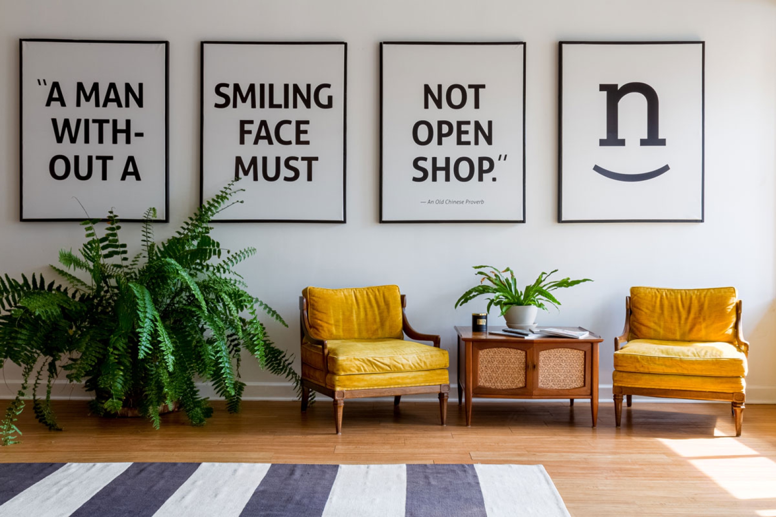 Photos by Kate Vogel Beautiful space with a large fern, two yellow upholstered lounge chairs, a nicely decorated coffee table and four-panels of large wall art reading: A man without a smiling face must not open shop.